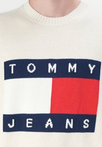 Maglione lavorato a maglia in crema con collo a giro. Caratterizzato da un audace logo blu navy, bianco e rosso che reca la scritta "TOMMY JEANS". Finitura testurizzata.