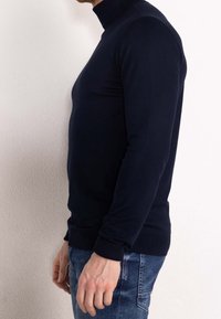 Granatowy, dopasowany sweter z golfem i długimi rękawami, z prążkowanym wykończeniem na dole i na mankietach, wykonany z gładkiej tkaniny. Noszony z jasnoniebieskimi, postarzanymi jeansami.