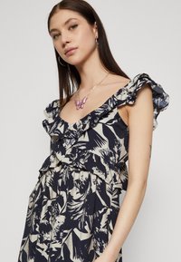 Robe fluide bleu marine et crème avec des bretelles à volants, motif floral et coupe décontractée. Portée avec un collier en forme de papillon rose et des boucles d'oreilles créoles.