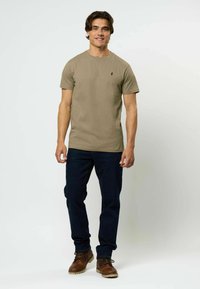 Chaki marškinėliai su apvaliu kaklu, trumpomis rankovėmis, ir mažas juodas emblema ant krūtinės, derinami su tamsiomis džinsomis ir rudais bateliais.