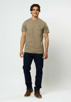 Khakifarbenes T-Shirt mit Rundhalsausschnitt, kurzen Ärmeln und einem kleinen schwarzen Emblem auf der Brust, kombiniert mit dunklen Jeans und braunen Stiefeln.