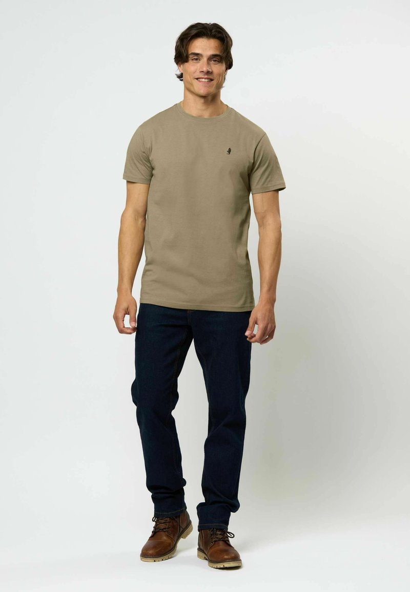 Chaki marškinėliai su apvaliu kaklu, trumpomis rankovėmis, ir mažas juodas emblema ant krūtinės, derinami su tamsiomis džinsomis ir rudais bateliais.