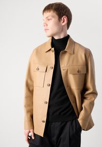 Νεαρός άνδρας που φοράει ένα καφέ σακάκι με κουμπιά πάνω από μαύρο ζιβάγκο, με το ένα χέρι στην τσέπη, κοιτάζοντας sideways σε ένα απλό ανοιχτό φόντο.