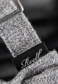 Correa de tela texturizada gris con hebilla de metal y una etiqueta negra con texto en cursiva blanca. El primer plano resalta el material y la costura.