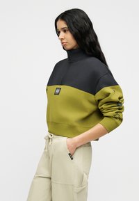 Γυναίκα που φοράει δίχρωμο φούτερ με ένα τέταρτο φερμουάρ σε μαύρο και ελιά, με γραφικό στο μανίκι, μπεζ παντελόνι με κορδόνι, με τα χέρια στις τσέπες, κοιτάζοντας προς τα αριστερά.