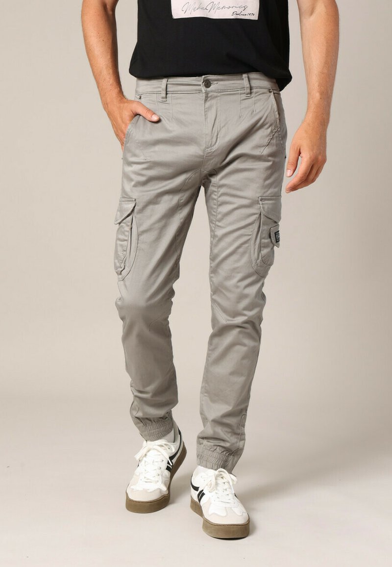 Man in lichtgrijze cargo broek met elastische boorden en witte sneakers, staand met één hand in zijn zak.