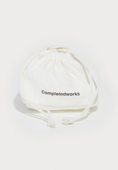 Bijela torba sviđača napravljena od mekanog materijala, sa crnim natpisom "Completedworks". Torba ima skupljeni vrh s podesivim vezicama.
