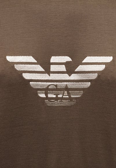 Καφέ μπλουζάκι από ύφασμα με λευκό κεντημένο λογότυπο που απεικονίζει στυλιζαρισμένα φτερά και το γράμμα "GA" στο κέντρο, παρουσιάζοντας μια ομαλή υφή.