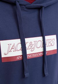 Marineblå hættekappe med et hvidt og burgunder logo med teksten "JACK & JONES" og "SINCE NINETEEN NINETY," struktureret stof og snore.