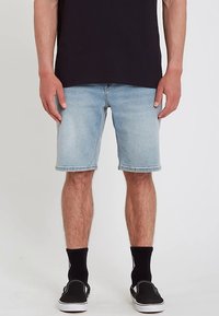 Shorts en jean bleu clair avec une coupe droite, ourlet effiloché et design à cinq poches. Portés avec des chaussettes noires et des chaussures sans lacets.