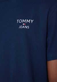 Camiseta de manga corta azul marino hecha de algodón, con un logo bordado en blanco "TOMMY JEANS" en la parte izquierda del pecho. Textura suave.