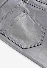 Silverfärgad metallisk tyg visar en närbild av en ficka, med en hexagonal form och framträdande sömnadsdetaljer. Slät yta är tydlig.