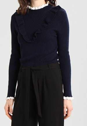 Mujer que lleva un suéter azul marino acanalado con volantes y puños blancos, combinado con pantalones negros de talle alto con cinturón de tela.