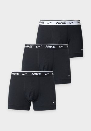 Skjorteløs mand med mørkt hår iført sorte Nike boxer briefs, stående barfodet mod en ensfarvet hvid baggrund med hænderne bag ryggen.