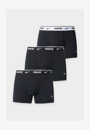 Särgita mees tumedate juustega, kandes musti Nike bokserpükse, paljajalu valge ühevärvilise tausta ees, käed selja taga.
