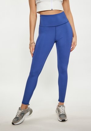 Kobieta ubrana w niebieskie legginsy z wysokim stanem i szare buty sportowe stoi z nogami lekko rozstawionymi na jednolitym tle.