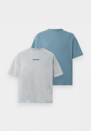 To modeller står side om side iført grå oversized t-shirts; den ene i sorte denimshorts og sorte sko, den anden i lyseblå jeans og sorte sko.