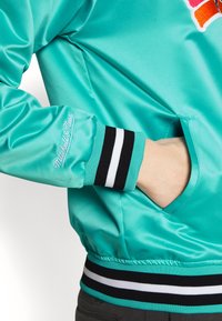 Chaqueta bomber de satén en un brillante color turquesa con franjas acanaladas en negro y blanco en los puños y el dobladillo; presenta un logo bordado en la manga.