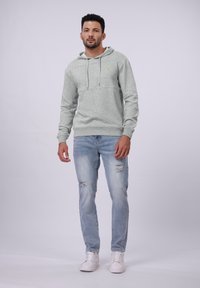 Sudadera gris con un acabado texturizado, acentos de diseño en ángulo, capucha con cordones, combinada con jeans claros desgastados y zapatillas blancas.