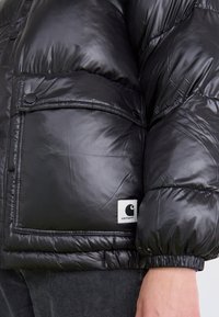 Čierna puffer bunda s lesklou prešívanou textúrou, uzatváraním na cvoky, veľkou prednou kapsou a výraznou bielou logom nášivkou.