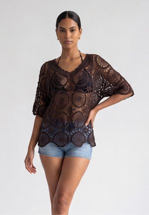 Donna che indossa un top nero in crochet trasparente sopra un top bikini nero, abbinato a pantaloncini di denim azzurri, in piedi con una mano sul fianco.