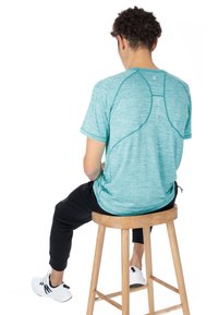 Turkos kortärmad träningsskjorta med ett texturerat mönster och kontrasterande sömmar. Svarta tapered byxor och vita träningsskor bärs.