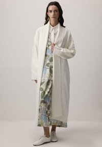 Dlhý krémový kardigán vyrobený z jemnej látky, s preloženým golierom a rebrovanými manžetami, pres doplnený kvetinovým šaty v modro-zelených odtieňoch.
