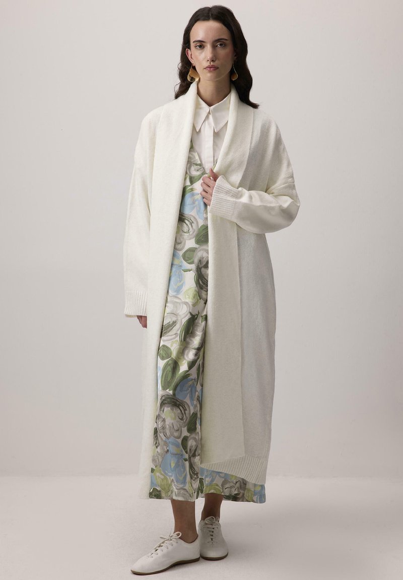 Dlhý krémový kardigán vyrobený z jemnej látky, s preloženým golierom a rebrovanými manžetami, pres doplnený kvetinovým šaty v modro-zelených odtieňoch.
