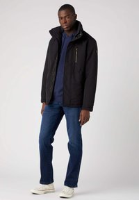 Czarna kurtka z wysokim kołnierzem i kieszeniami na zamek, noszona na niebieskiej bluzie z kapturem, połączona z niebieskimi dżinsami i białymi sneakersami. Gładka tekstura.