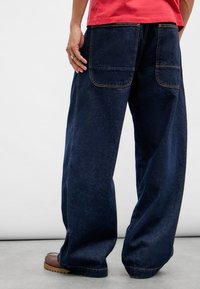 Mørkeblå denim brede bukser med to baklommer, forsterkede s�ømmer og kontrasterende beige sting, stylet med en rød skjorte.