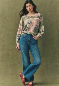 Gebreide trui met multicolored landschappatroon, lichte roze en groene kleuren; gecombineerd met brede blauwe denim jeans en rode platte schoenen.