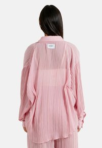 Blusa rosa claro con textura plisada, puños abotonados y cuello doblado. Presenta una pequeña etiqueta en la parte superior de la espalda.