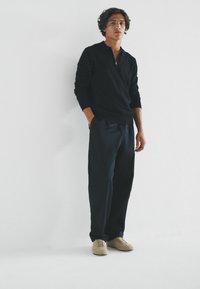 Černý svetr na zip s dlouhými rukávy, doplněný tmavě námořnickými kalhotami a světlými béžovými semišovými botami. Neutrální pozadí zdůrazňuje outfit.