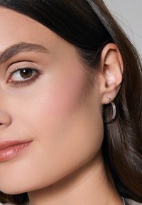 Srebrny okrągły kolczyk na uchu modelki, o fakturowanym wykończeniu. Odcień skóry modelki jest ciepły, a tło jest neutralne i delikatnie rozmyte.