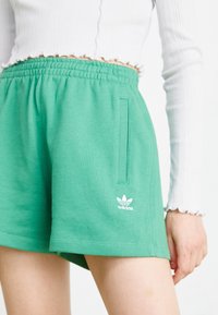 Žena obléká zelené šortky Adidas s elastickým pasem a boční kapsou, spárované s bílým dlouhým ribbovým topem s krátkým rukávem.