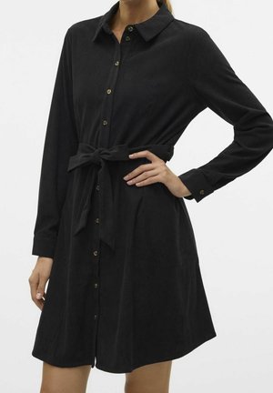 Frau trägt ein schwarzes, langärmliges, knöpfbares Kleid mit einem gebundenen Stoffgürtel in der Taille, steht mit einer Hand auf der Hüfte.