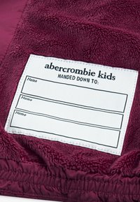 Material de fleece de culoare burgundy cu o etichetă albă pentru nume. Eticheta scrie "abercrombie kids" și include trei câmpuri goale pentru nume.