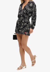 Vestido negro con estampado de paisley, mangas largas, cintura ceñida y escote en V. Combinado con sandalias blancas de tiras y un pequeño bolso negro.