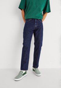 Mörkblå jeans med rak passform, kombinerat med en grön kortärmad skjorta och gröna och vita rutig slip-on skor.