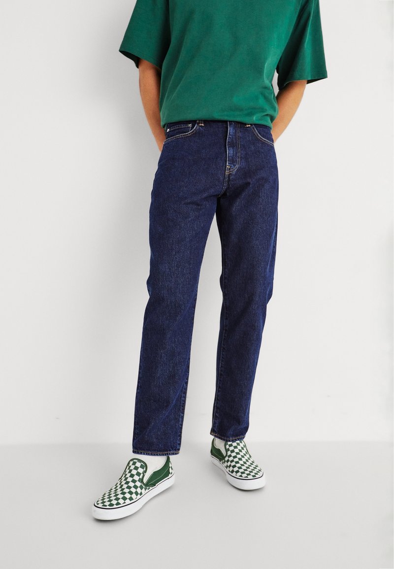 Mörkblå jeans med rak passform, kombinerat med en grön kortärmad skjorta och gröna och vita rutig slip-on skor.