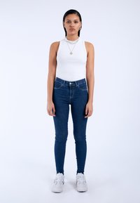 White ribbed tank top, dark blue skinny jeans with front pocket detail, white athletic shoes, and layered necklace. Plain background.