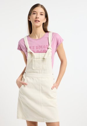 Crème corduroy tuinbroekjurk met verstelbare banden, een voorzak en zijknopen; gecombineerd met een roze graphic T-shirt.