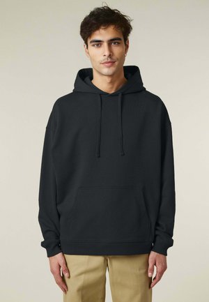 Jonge man in een zwarte hoodie met een voorzak en trekkoorden, gecombineerd met beige broek, staand tegen een eenvoudige lichte achtergrond.