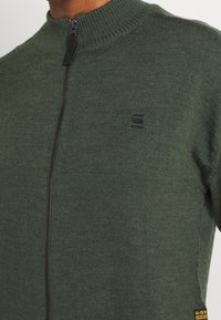 Groene zip-up trui met hoge kraag, van textuurgebreid materiaal, met een klein logo op de borst en een zwarte rits.