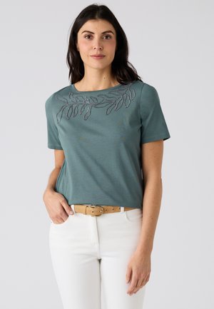 Mujer con una camiseta de manga corta de color azul verdoso con un diseño bordado de hoja cerca del escote, pantalones blancos y cinturón marrón claro, posando frente a un fondo liso.