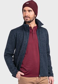 Marineblaue, strukturierte Jacke mit Stehkragen, Reißverschluss und zwei seitlichen Taschen, getragen über einem bordeauxfarbenen Henley-Shirt. Braune Hose rundet das Outfit ab.