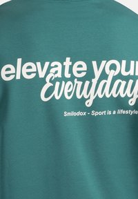 Camiseta de cuello redondo en color azul verdoso, hecha de algodón. Presenta texto blanco que dice "eleva tu día a día" y "Smilodox - El deporte es un estilo de vida" en la parte posterior.