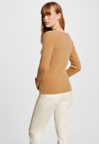 Maglione marrone dalla texture particolare con un design aderente e maniche lunghe, caratterizzato da bottoni sui polsini, abbinato a pantaloni color crema chiara.