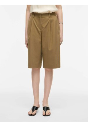 Nő, aki barna, térdig érő szabott rövidnadrágot, fehér felsőt, fekete lapos, nyitott orrú szandált, ezüst karkötőt és gyűrűket visel, fehér padlón áll.