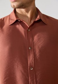 Chemise à boutons dans un orange brûlé, tissu léger avec une texture lisse. Présente un col évasé et deux boutons à l'avant.